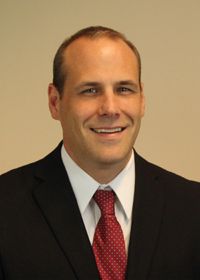 Professional headshot of Jeff Heinecke, wearing a black suit, white shirt, and red tie, smiling against a neutral background.