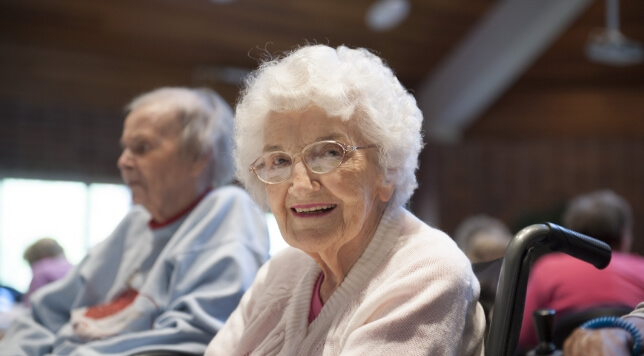 Elderly woman with white hair and glasses smiling while sitting in a wheelchair.