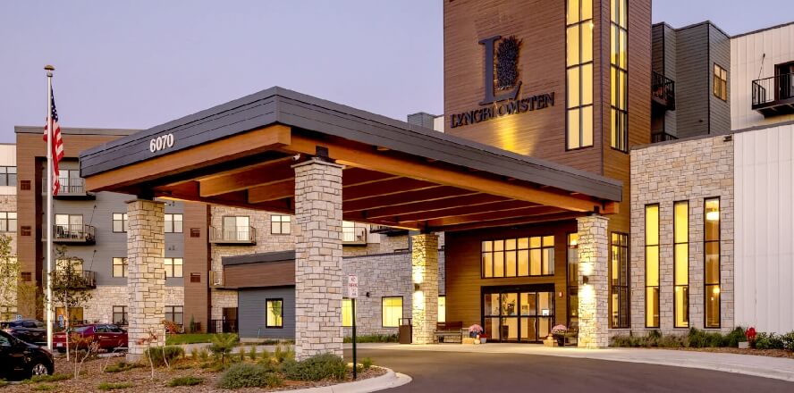 Entrance of a modern senior living facility with a covered driveway, stone pillars, and illuminated windows at dusk.