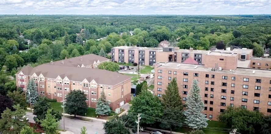 Aerial view of a senior living campus surrounded by greenery, featuring multiple brick buildings and landscaped grounds.