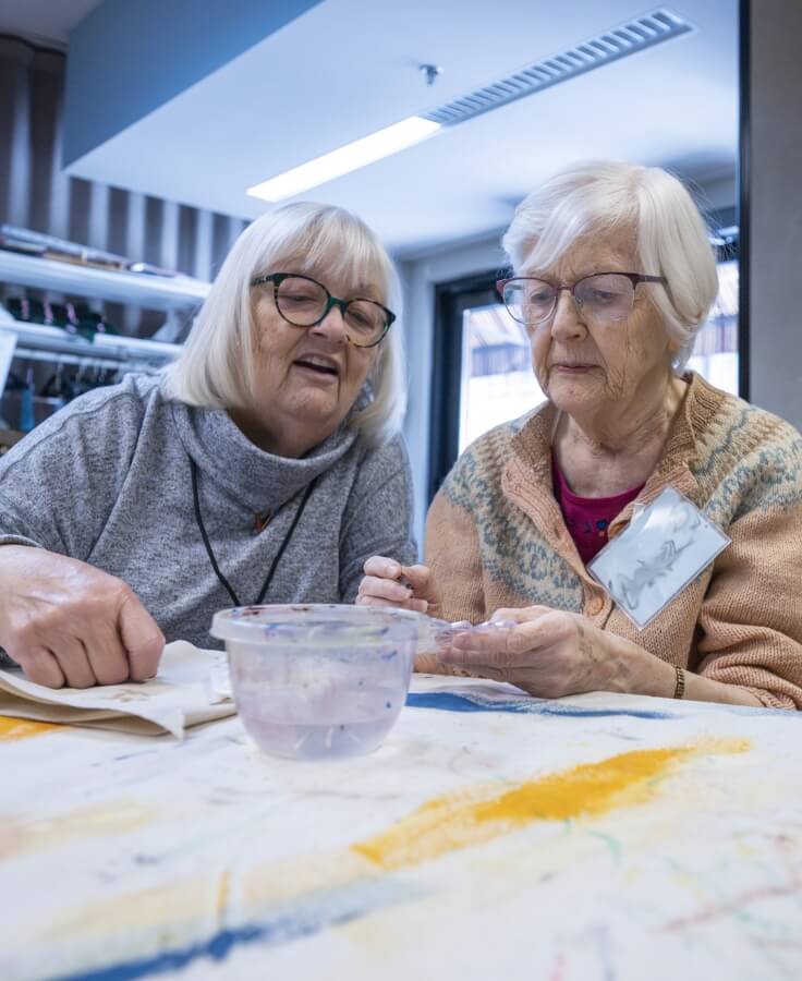 Two elderly women with glasses working together on an art project, one holding a small object while the other looks on with a smile.