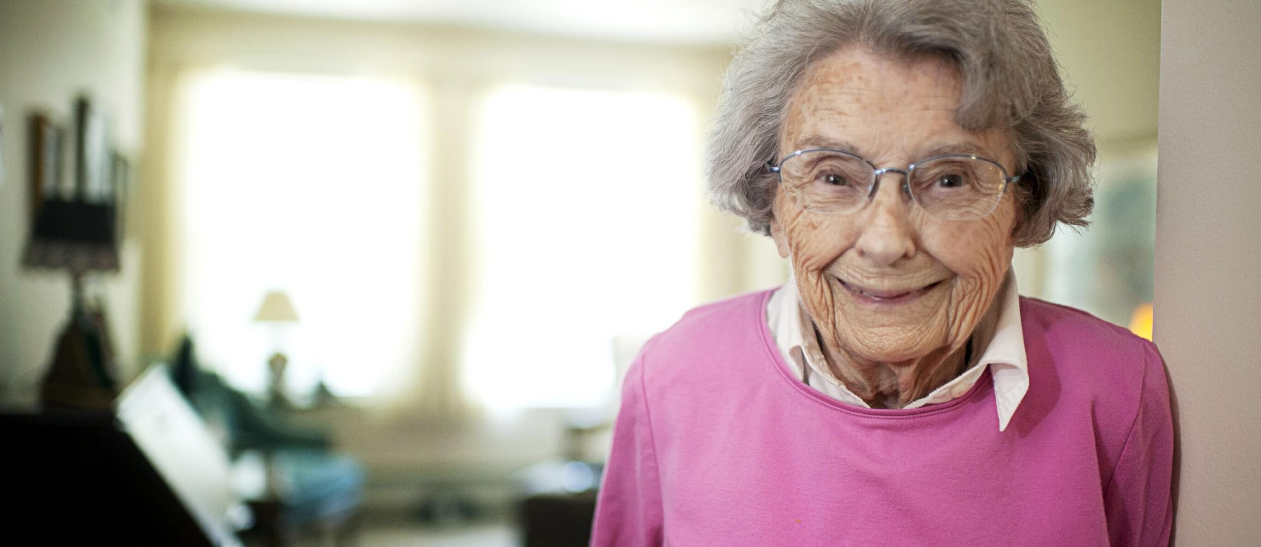 Smiling older woman wearing glasses and a pink sweater in a bright living room.
