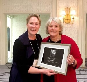 Two smiling women hold a framed award certificate at a formal event, standing in a well-lit room with elegant decor.