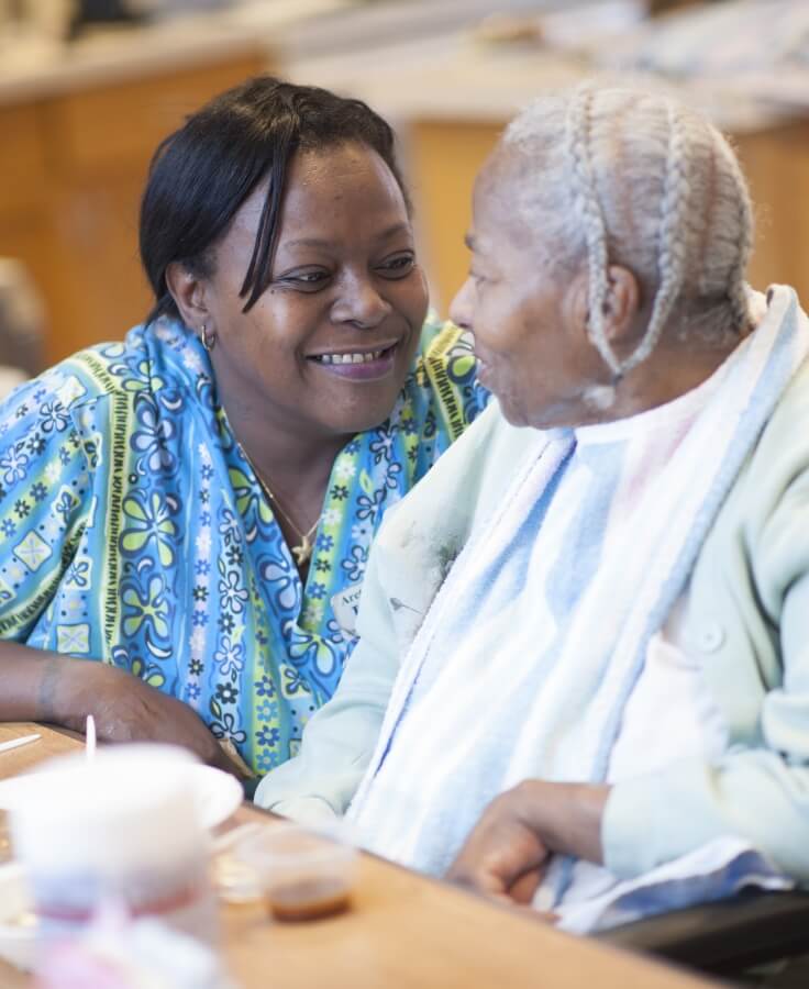 A smiling caregiver in a blue and green patterned uniform leans in to engage warmly with an elderly resident in a wheelchair, who is wearing a bib.