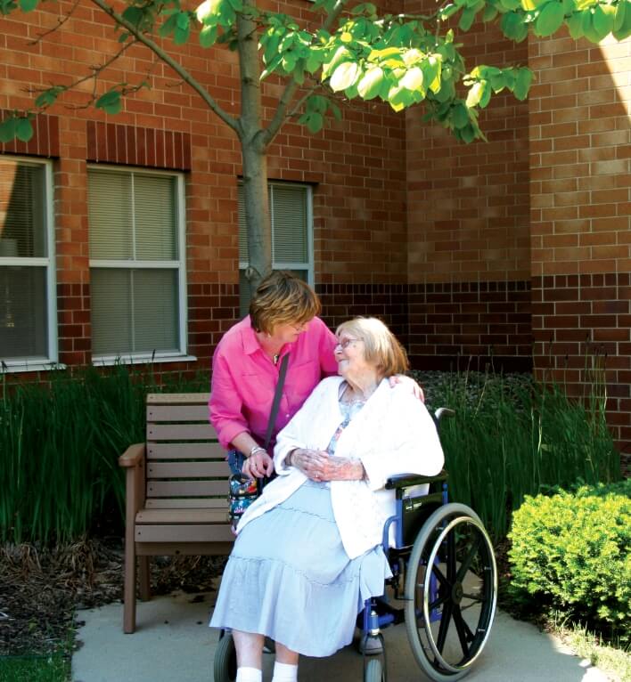 A caregiver in a pink shirt leaning towards an elderly woman in a wheelchair, sharing a warm moment outdoors.