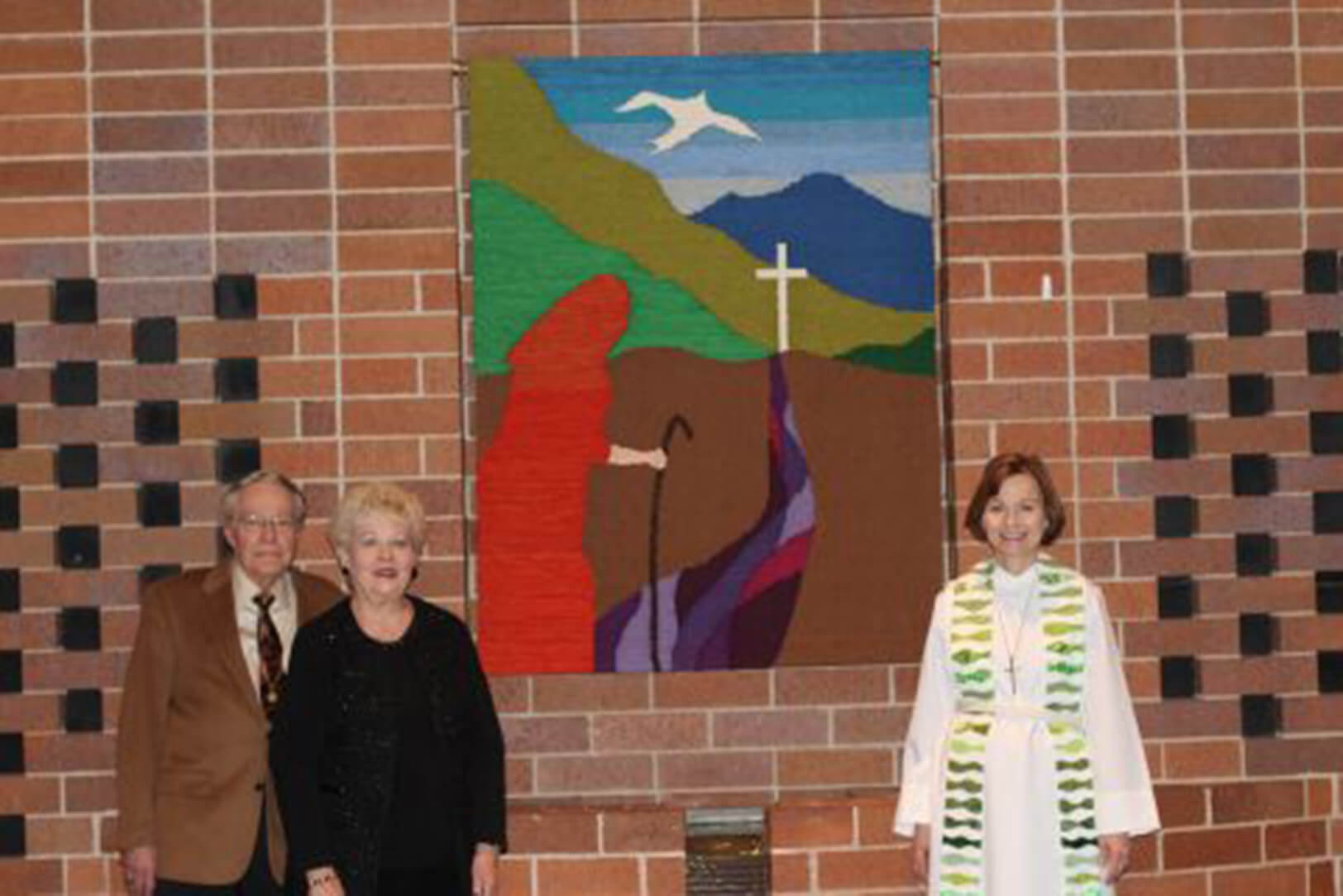 Two elderly individuals and a clergy member in a white robe with green accents stand in front of a colorful religious tapestry depicting a cross, a dove, and a robed figure.