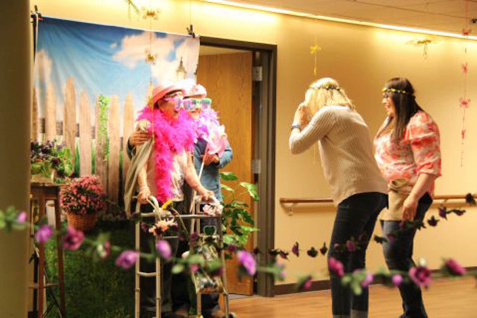 A group of people, including a senior using a walker, dressed in colorful costumes and accessories, pose for a fun photo booth session with a cheerful outdoor-themed backdrop.