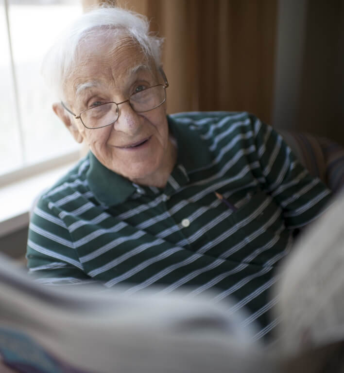 A smiling elderly man with glasses and a green striped polo shirt sits near a window, holding and reading a newspaper.