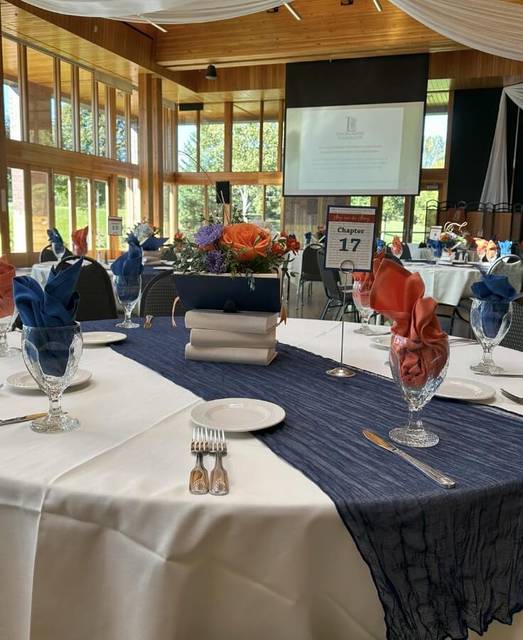 A beautifully decorated banquet table set for an event, featuring a navy blue table runner, folded napkins, glassware, and a floral centerpiece atop a stack of books.