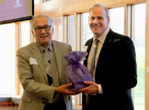 Two men in formal attire smiling as they exchange a gift wrapped in purple fabric at a professional gathering.