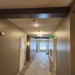 A hallway under renovation with protective floor covering, modern lighting, and an exit door leading to natural light.