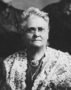 A black and white historical portrait of a woman wearing glasses, a lace shawl, and a decorative brooch, with a composed expression.