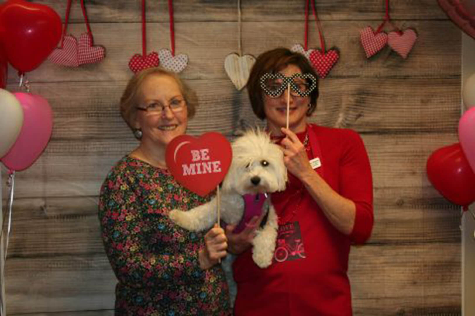 Two smiling women pose with a small white dog at a Valentine's Day-themed photo booth, holding heart props and wearing festive accessories.