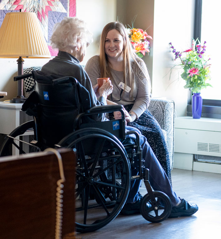 Therapeutic Recreation Coordinator sits and chats with a care center resident as she sips her coffee in her room.