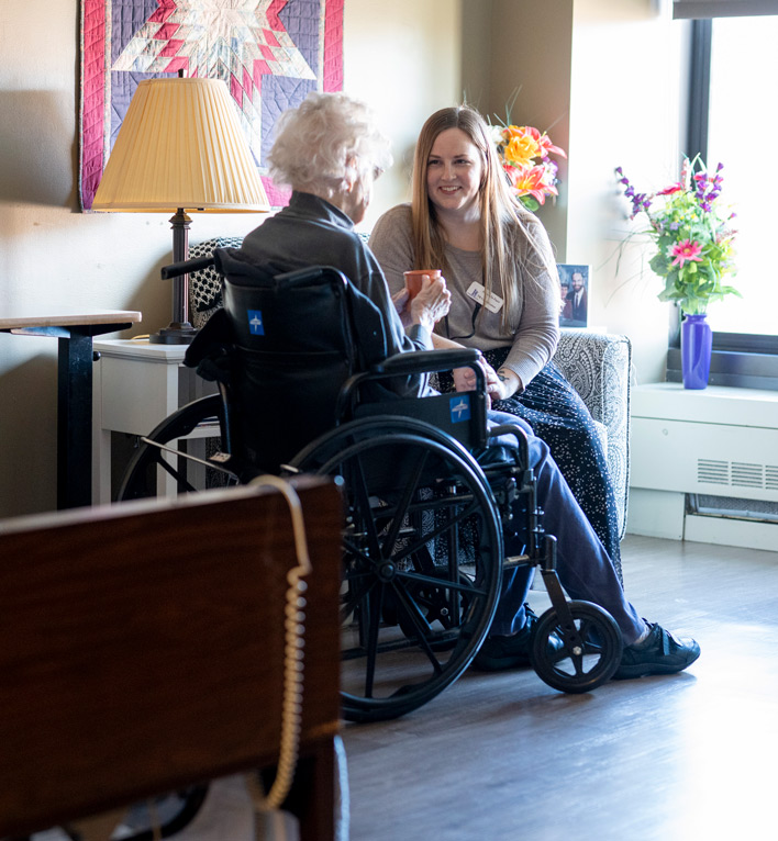 Therapeutic Recreation Coordinator sits and chats with a care center resident as she sips her coffee in her room.
