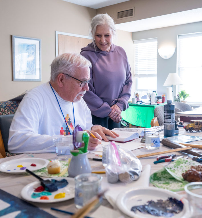 Art with Heart facilitator watches a resident continue his art project. The table is filled with lots of mediums: paint, watercolor, pencils, and much more.