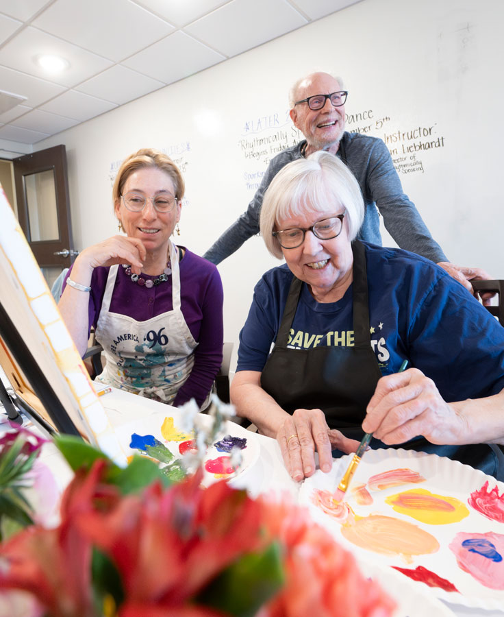 The teaching artist and another resident look over a resident's shoulder as she paints flowers with colorful acrylics on a canvas in the Studio.