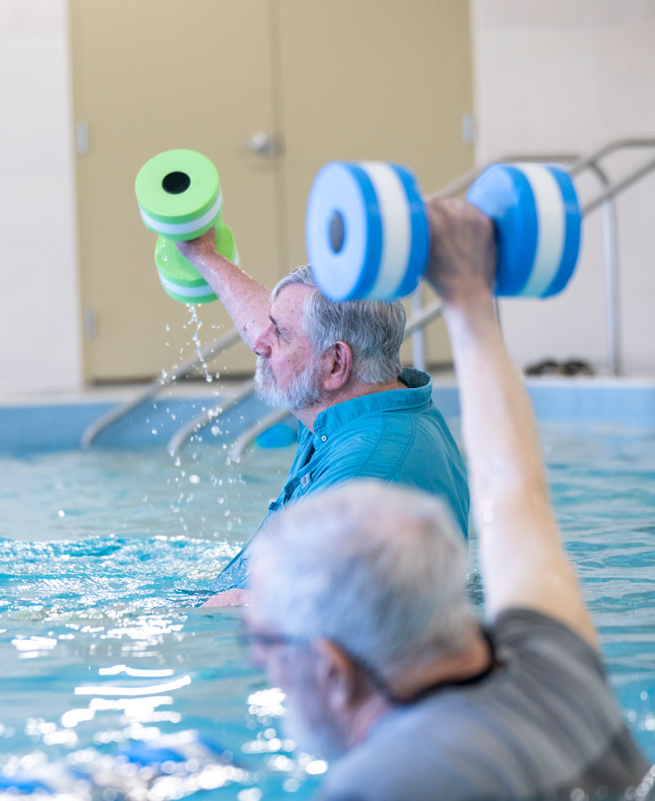 Residents attend an exercise class in the warm water pool. Pictured are two men lifting weights above their heads.