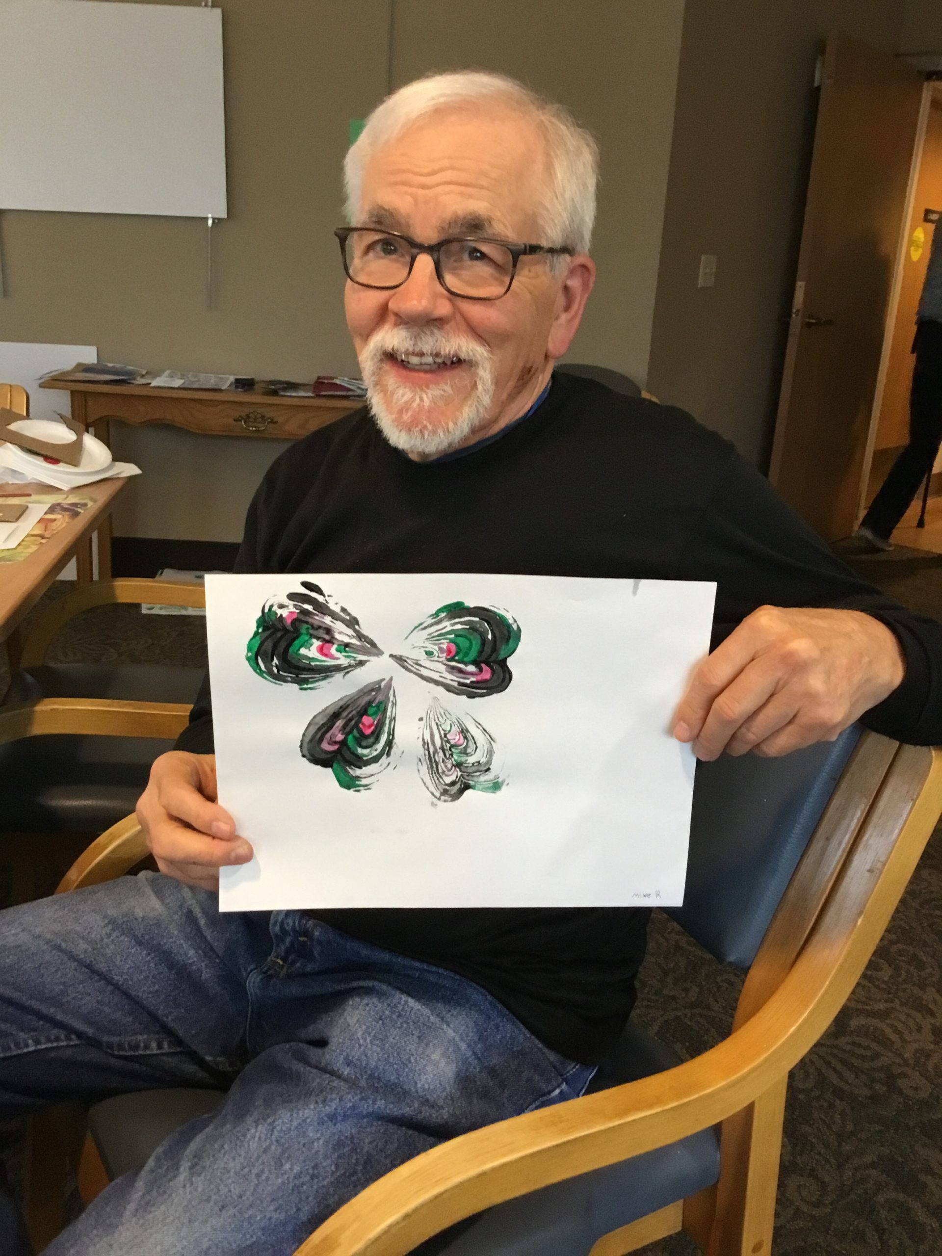 A resident from The Heritage at Lyngblomsten holds up art (colored black, pink, and green) resembling a clover while participating in Art with Heart.