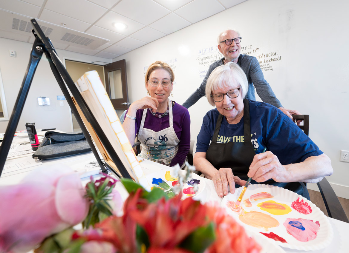 Teaching Artist and Resident Watch as another Resident Paints Flowers with Acrylic Paint in the Studio